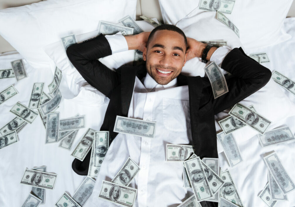 Happy African man in suit lying on bed in hotel room with falling money and looking at camera